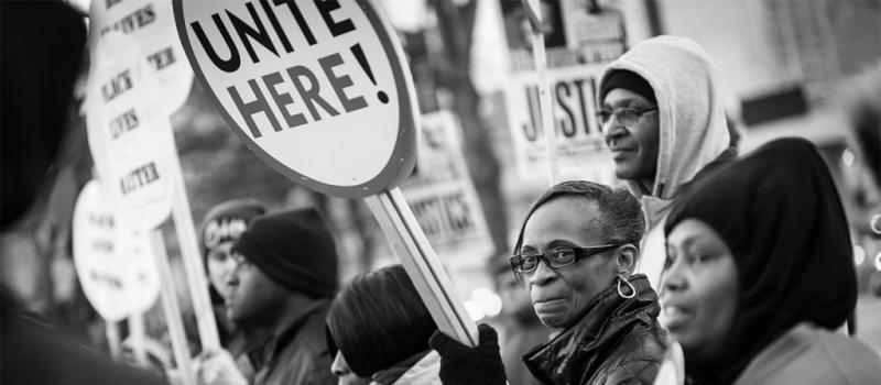 Photo of protestors holding signs. Image is focused on African American woman holding a sign that says, "Unite Here!"